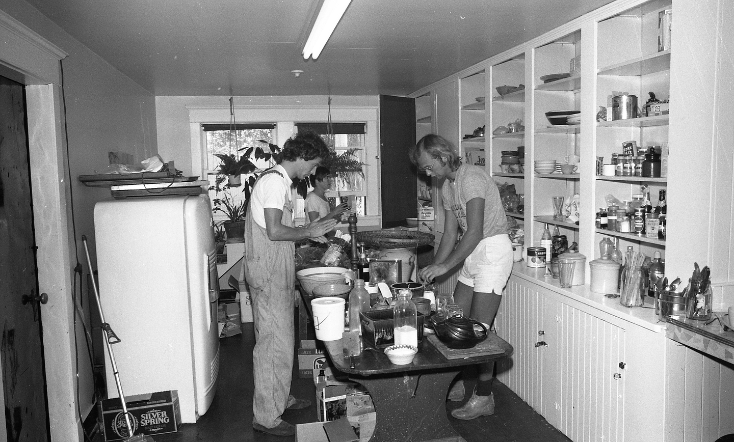 A black and white photograph of Warren Knechtel, Kate Craig and Vincent Trasov standing around a table in the kitchen, preparing food.