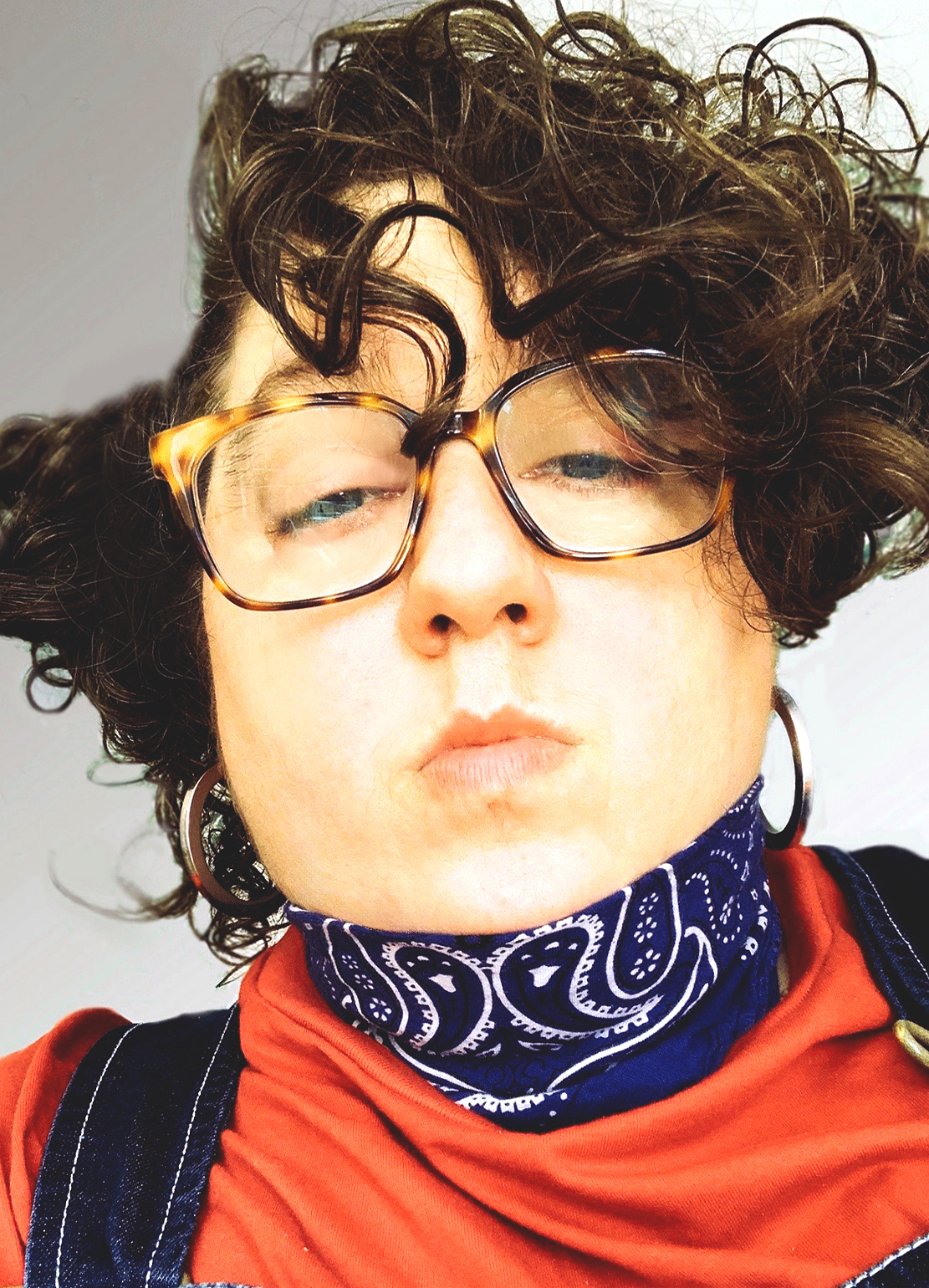 Close-up, low-angle selfie of Constantina Zavitsanos with tousled dark curly hair, large tortoiseshell glasses and silver hoop earrings, eyes half-closed and lips slightly pursed. They wear a navy paisley bandana knotted at the neck, an orange-red T-shirt and dark denim overalls, against a pale wall.