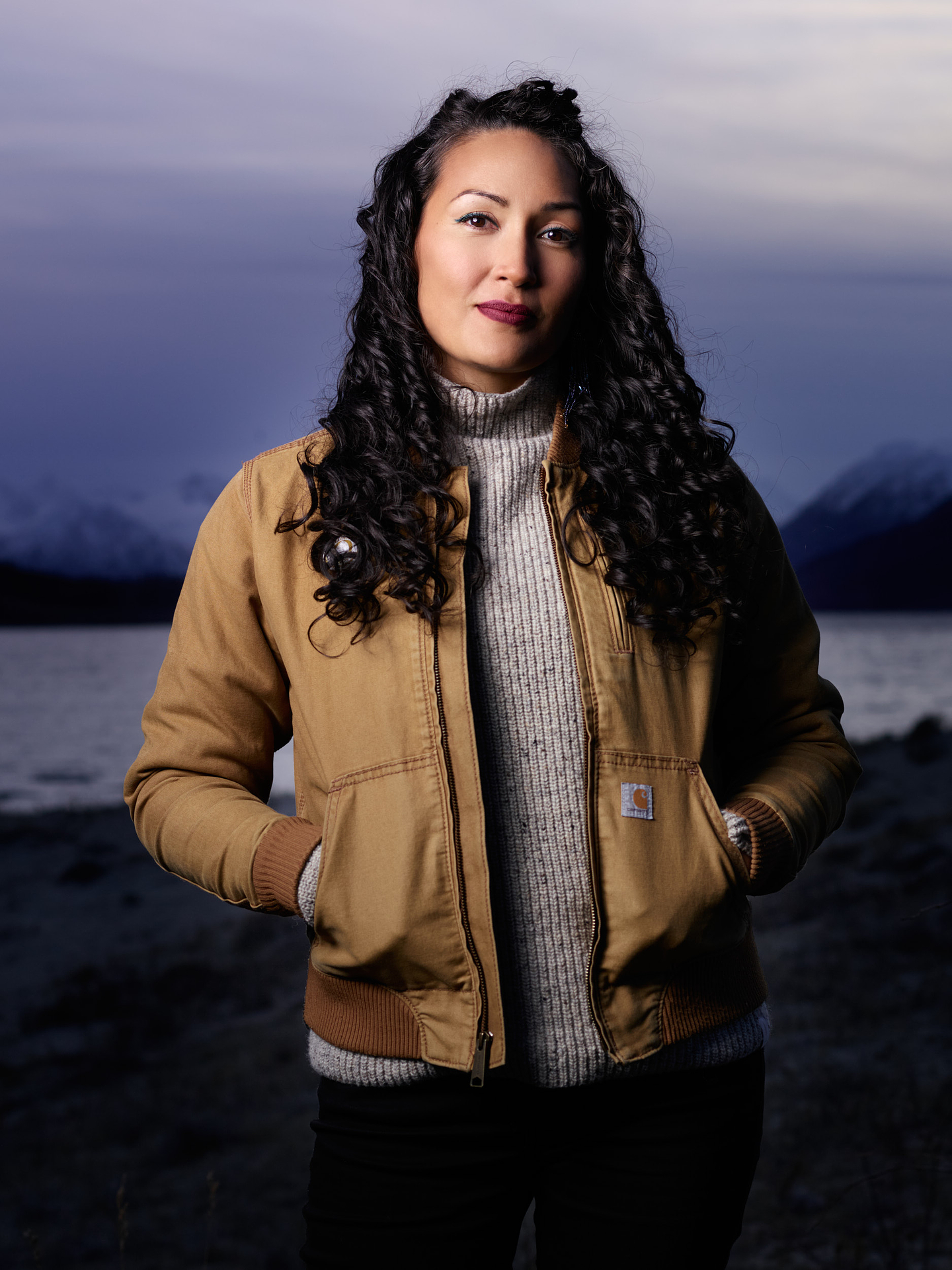 A portrait of Siku Allooloo looking directly at the camera with her hands in the pocket of her jacket. She is standing outside and a lake, snow capped mountains, and a mauve sunset can be seen in the background.