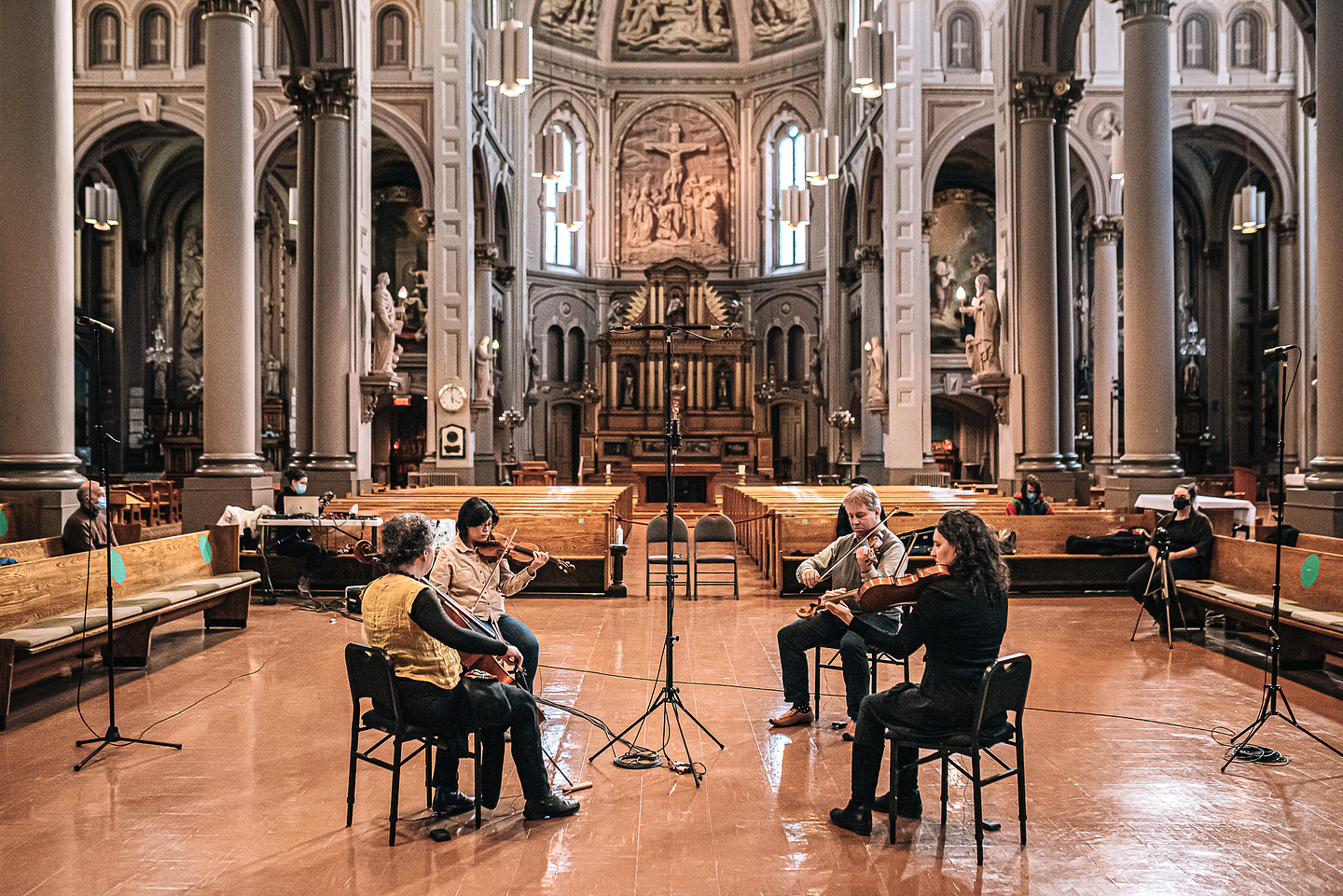 A string quartet playing their instruments in a church. They sit facing each other, with microphones placed in between and on each side of them. In the background, a few other people are sitting on the church pews in front of a background of columns, an organ, murals and a crucifix.