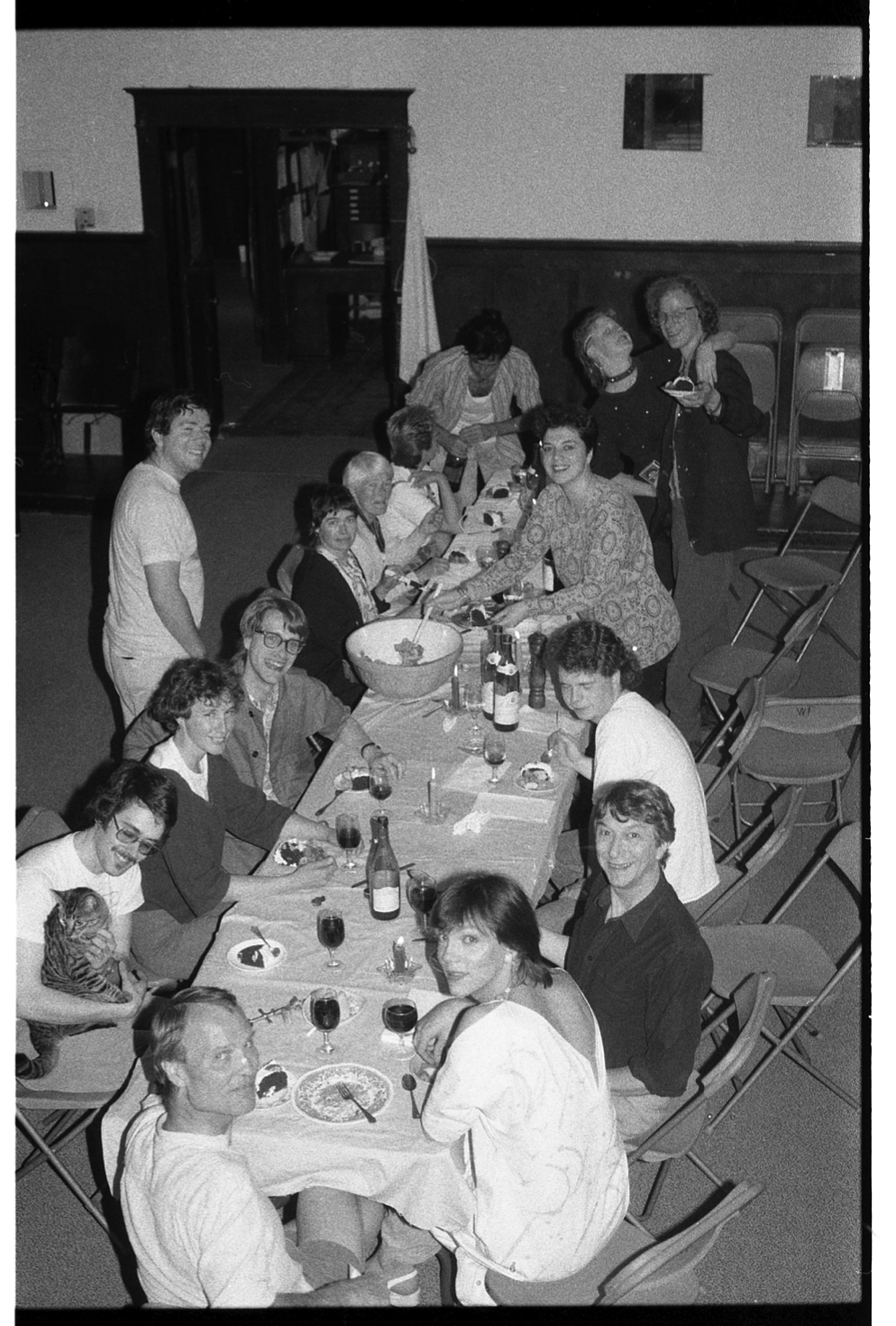 A black and white photograph of fifteen people seated around a long table in the Grand Luxe Hall, with glasses of wine and plates of cake. A tabby cat sits on one guest’s lap.