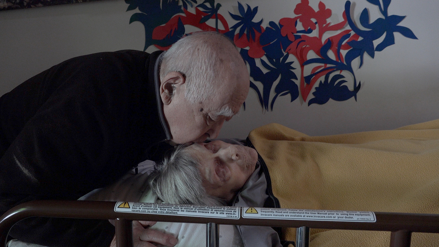 Nobuo Kubota's wife Lee lies in a hospital bed under a yellow blanket while Nubuo tenderly places a kiss on her forehead, in a dim room with colorful abstract wall art in the background.