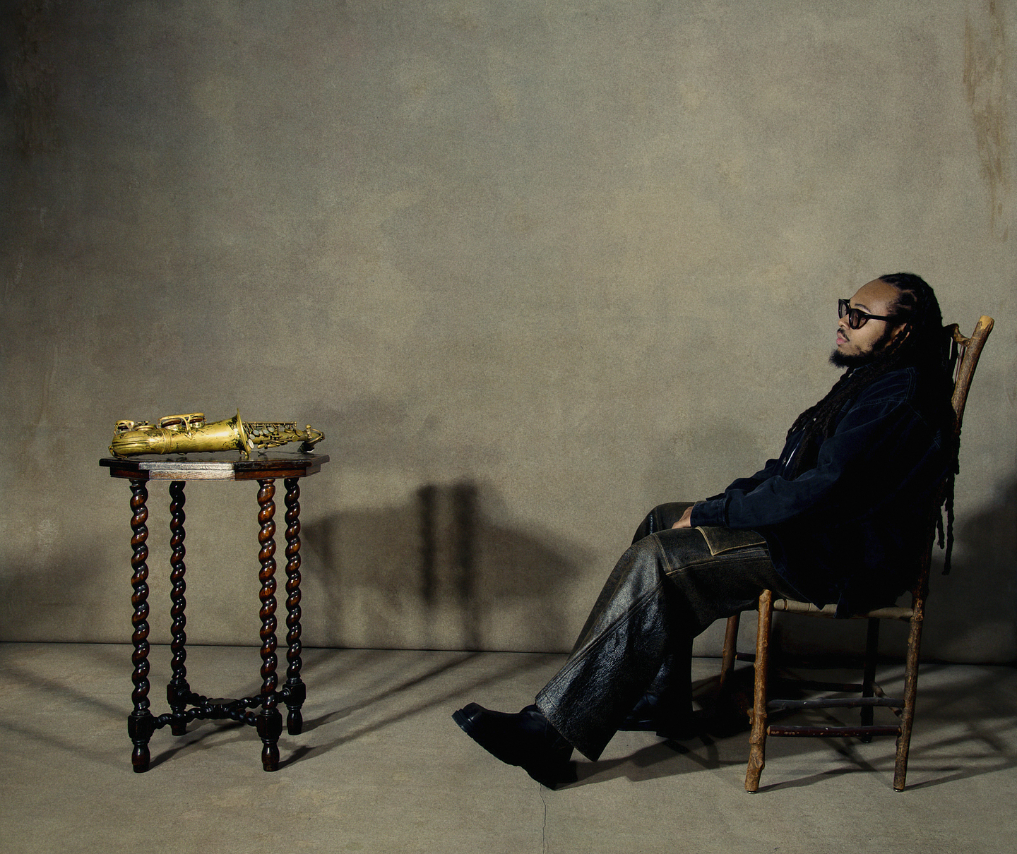 Immanuel Wilkins wears sunglasses as he sits in a wooden chair facing left in a minimalistic, dimly lit room. He wears a dark jacket and leather pants. To his left is a small ornate wooden table with a brass saxophone resting on it. The background is a plain, textured wall, casting soft shadows.