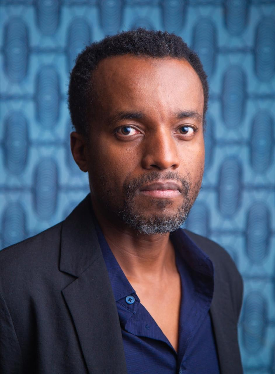 Portrait of Kivu Ruhorahoza with short dark hair and a greying beard, wearing a black blazer over an open-collared navy shirt and looking directly at the camera with a calm, steady expression. Behind him is a blue patterned backdrop of repeating oval, fingerprint-like motifs.