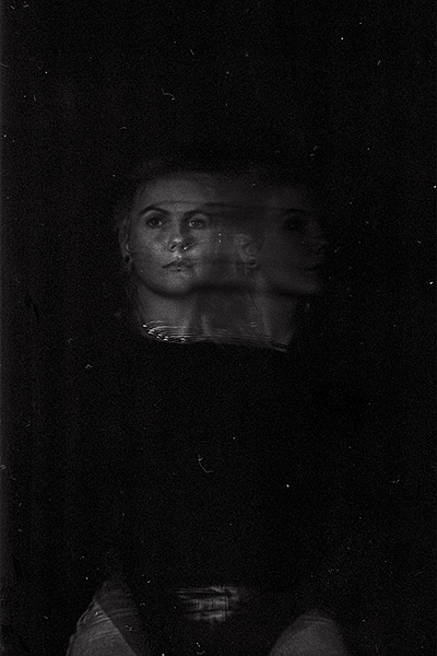 A black-and-white long exposure photo of Sidney Gordon. They gaze at the camera directly while a ghostly blur maps movement around their head. 