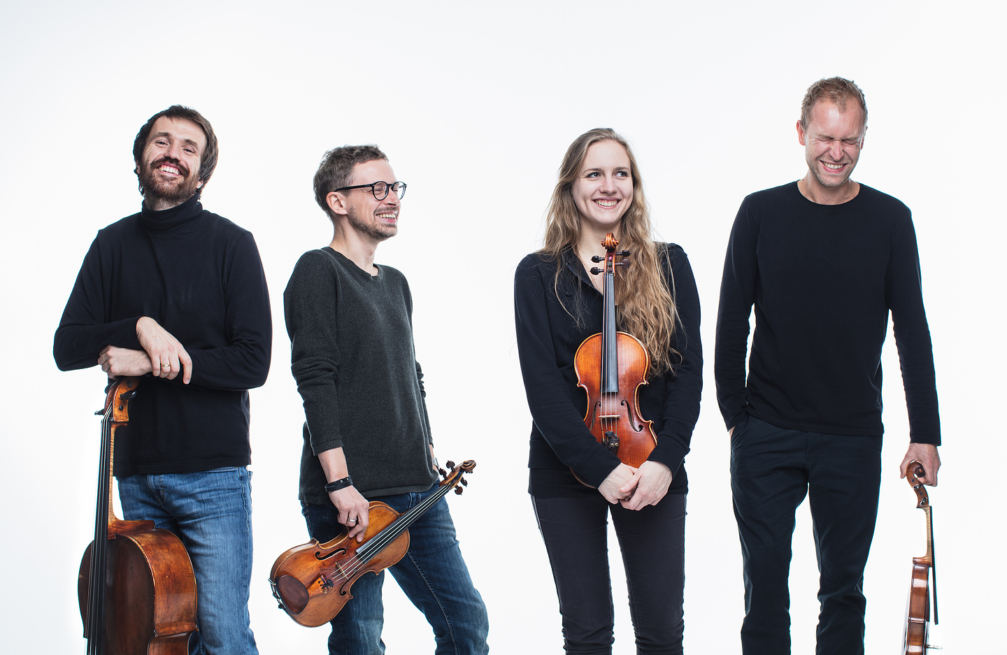 Four musicians from the Lutosławski Quartet stand against a white background, smiling and laughing. The first person on the left holds a cello, the second holds a viola, the third (a woman) holds a violin in front of her, and the fourth holds a violin by his side. They are dressed casually in dark tops and jeans or dark pants.