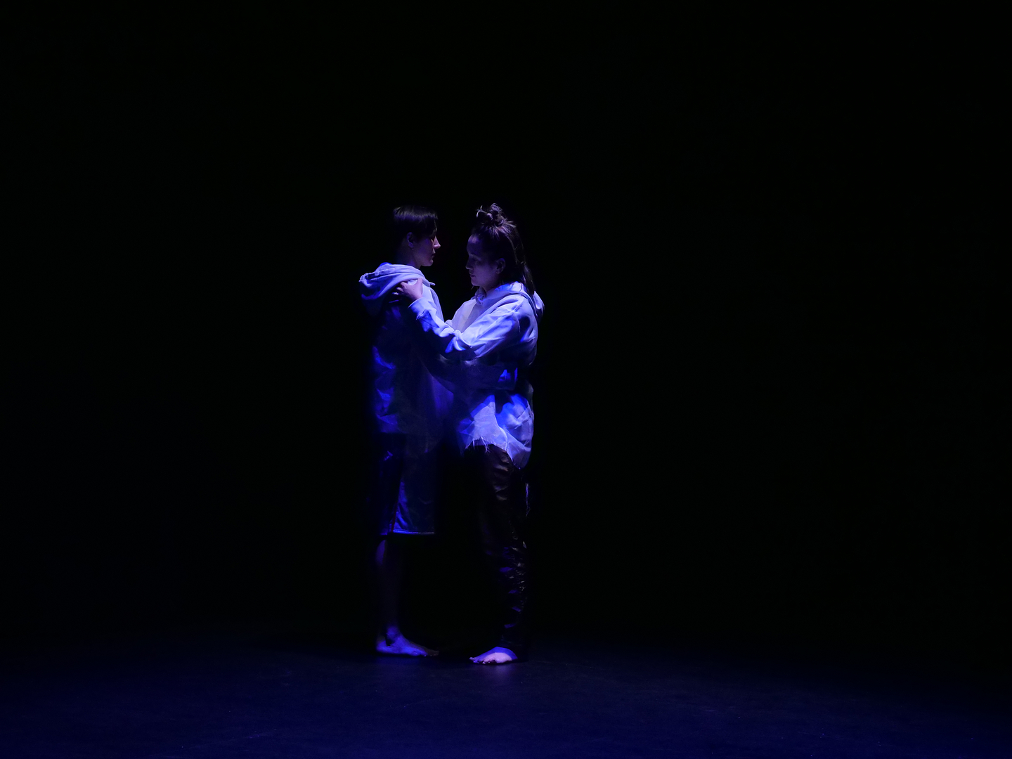 Two people stand facing each other in profile in a dark, purple space that looks like it goes on forever.  Their hands rest on each others’ shoulders. They wear white hooded sweatshirts. Their feet and legs are shadowed while their faces and torsos are illuminated by blue-white light. 