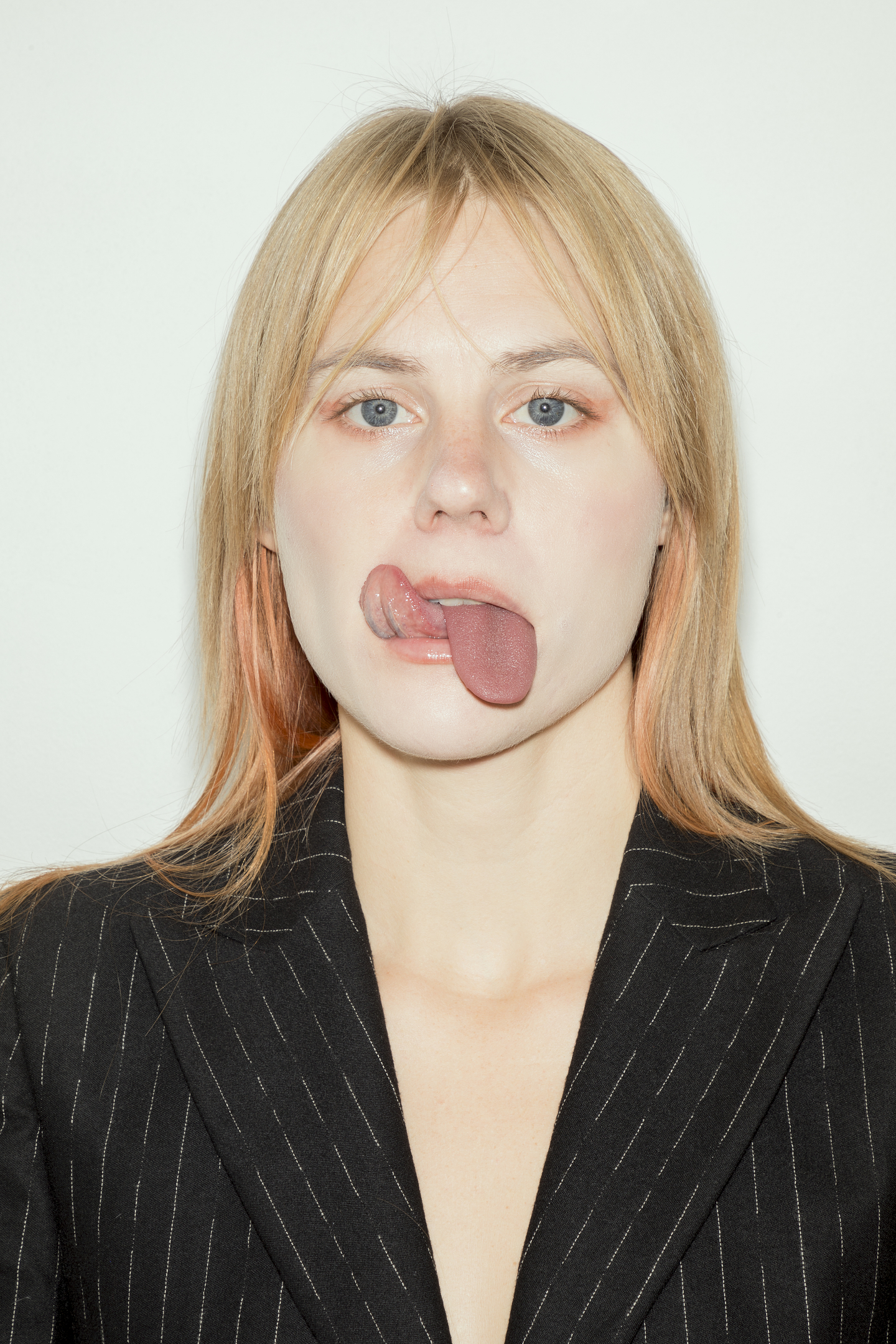 A headshot of Stina Fors. She stares directly at the camera with her tongue reaching to her upper lip, with a second prosthetic tongue dangling from her mouth. She has platinum blonde hair and wears a black pinstriped blazer. 