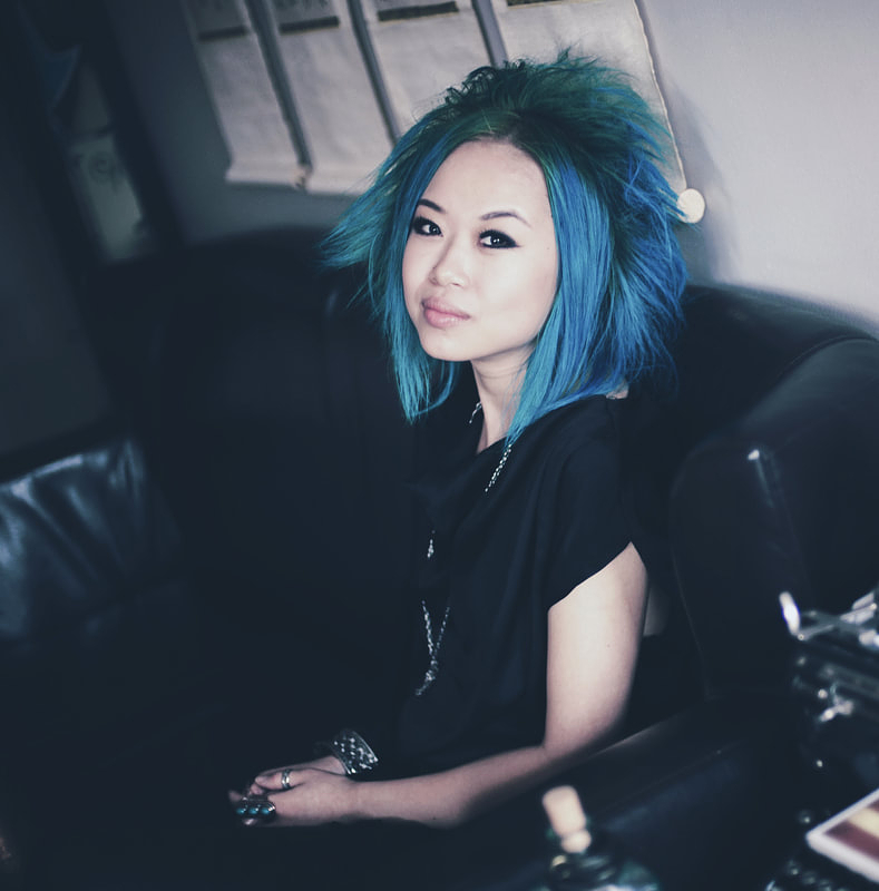 Jen Sungshine sits on a black vinyl couch and turns to look at the camera. She has bright blue hair, and wears a black t-shirt and silver jewellery.
