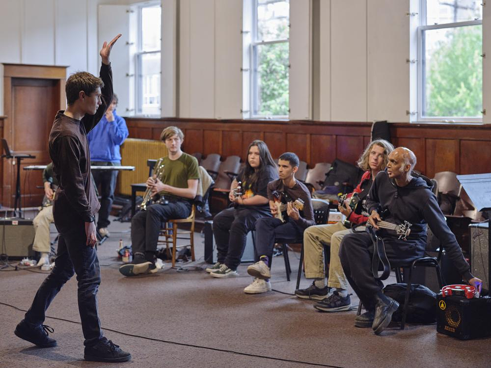 A young person with short-cropped brown hair stands with one arm raised to the ceiling, facing a row of musicians. They wear black pants and a brown long-sleeve shirt. The group features several guitarists, a pianist, and a brass musician. 