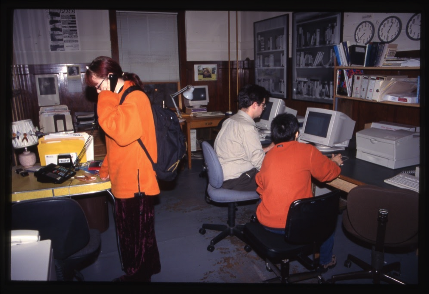 A colour photograph of Western Front staff in the office. Anna Stauffer stands next to a table holding a phone to her ear, and Jonathan Middleton and Eileen Kage are seated at a desk, looking at a computer.