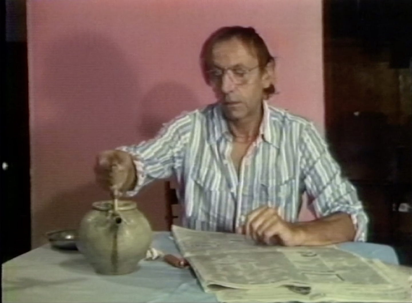 A man wearing a blue and white striped shirt sits at a table. With his right hand he reaches for a ceramic teapot, while his left hand rests on a newspaper. Behind him is a bright pink backdrop.