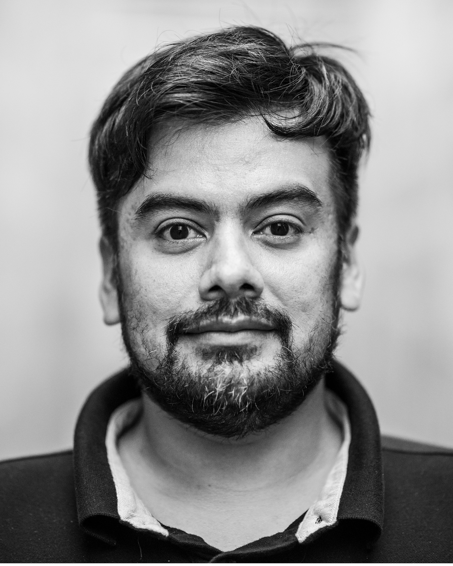 Black-and-white portrait of Naufus Ramírez-Figueroa with tousled dark hair and a beard, wearing a dark collared shirt over a light undershirt and looking directly at the camera with a slight smile. The background is a soft, out-of-focus pale wall.
