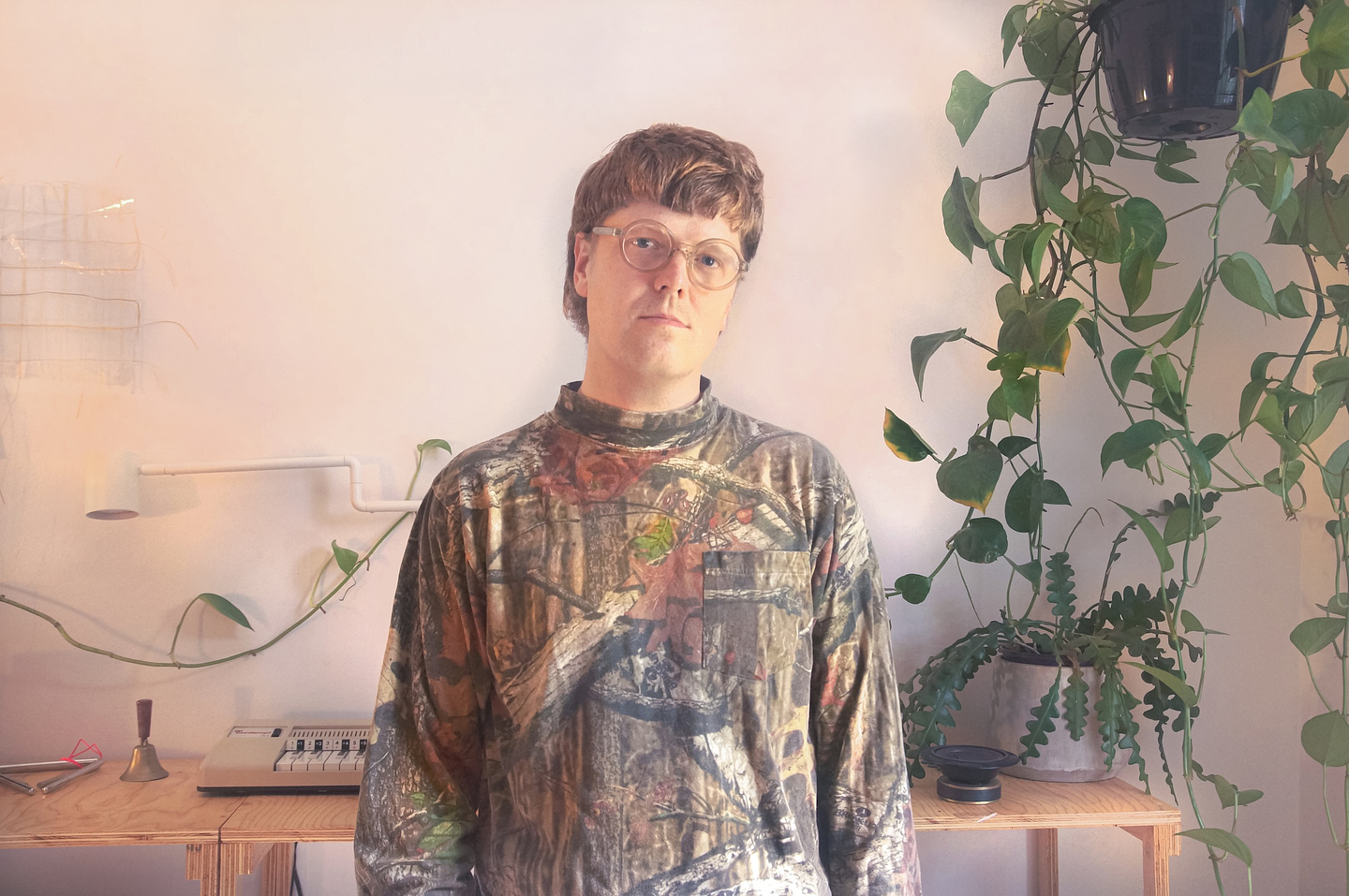 Portrait of a person wearing glasses and a patterned shirt, standing in a softly lit room beside a table with audio equipment and several trailing houseplants.