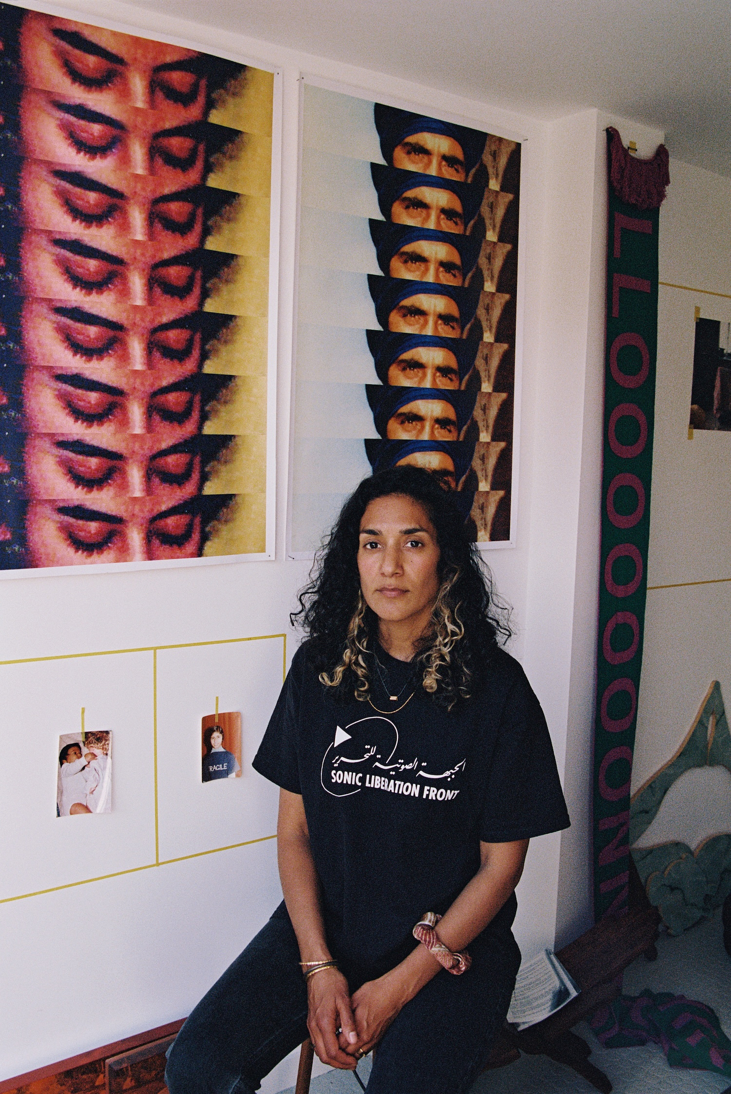 A portrait of Jasleen Kaur with long, dark curly hair with blonde highlights, seated on a stool in a white studio and looking directly at the camera with a calm expression. She wears a black T-shirt printed with "Sonic Liberation Front" in English and Arabic, gold necklaces and bracelets, and a patterned fabric wristband. Behind them hang two large prints, one repeating a close-up of heavily lined eyes in a vertical strip and the other repeating an image of a man in a blue turban. Small snapshots are taped inside yellow tape outlines on the wall, and a long green-and-pink knitted banner reading "LLOOOOOOON" hangs at the right.
