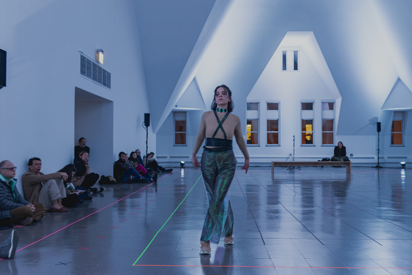 Nour Mobarak walks across a gallery floor with her eyes closed and arms outstretched behind her. She wears a glowing choker, a sheer, long-sleeved beige crop top, a black X-shaped brace across her chest, iridescent pants, and sneakers. Lines of neon green and pink tape demarcate the performance space, and audience members sit on the periphery against the gallery wall. The back wall of the space is architecturally distinct, as it features a row of windows below dramatic angular recesses. 