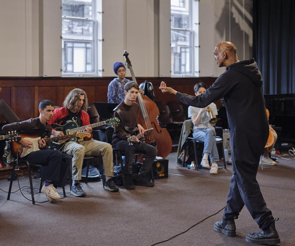 A group of young musicians sit in a row, facing a bald instructor wearing all black. The group features several string instrumentalists and a brass musician. The instructor gestures at the musicians with one hand, stepping towards the group. 
