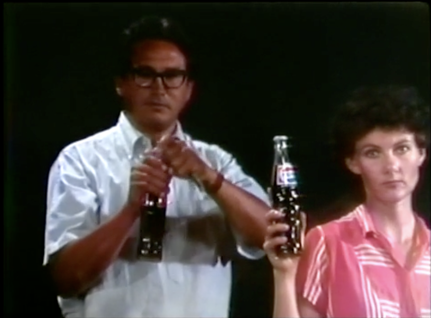 A man in a white shirt opens a bottle of Coca Cola, while a women in a pink dress holds up a bottle of Pepsi with a look of disdain on her face. 