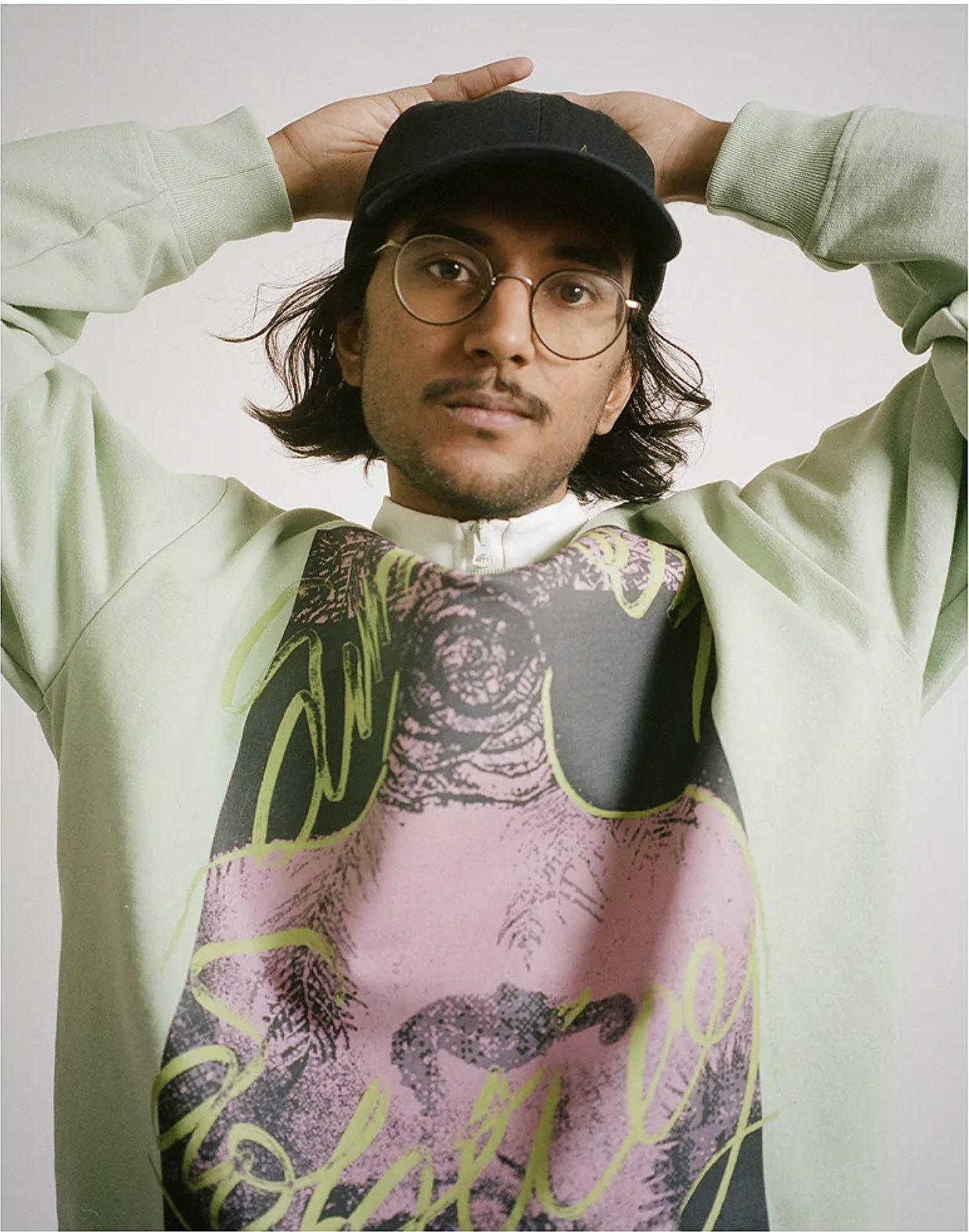 Portrait of a person wearing round glasses, a black cap, and a pale green graphic sweatshirt, with their hands resting on top of their cap against a plain light background.