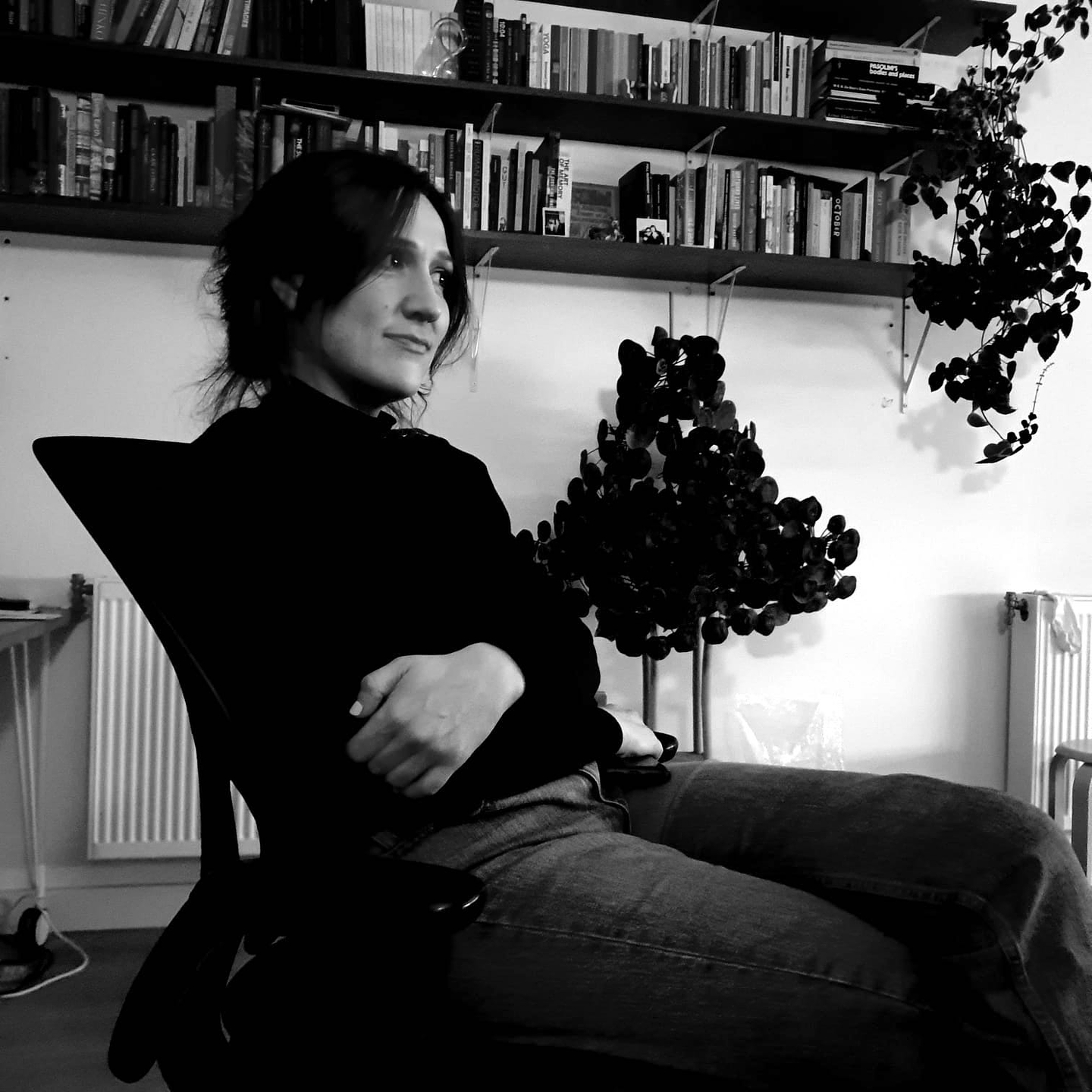 A black and white photo of Holly Márie Parnell sitting in a chair in a domestic interior. She folds her arms and smiles shyly while looking to the side. Behind her are shelves of books and a number of plants.