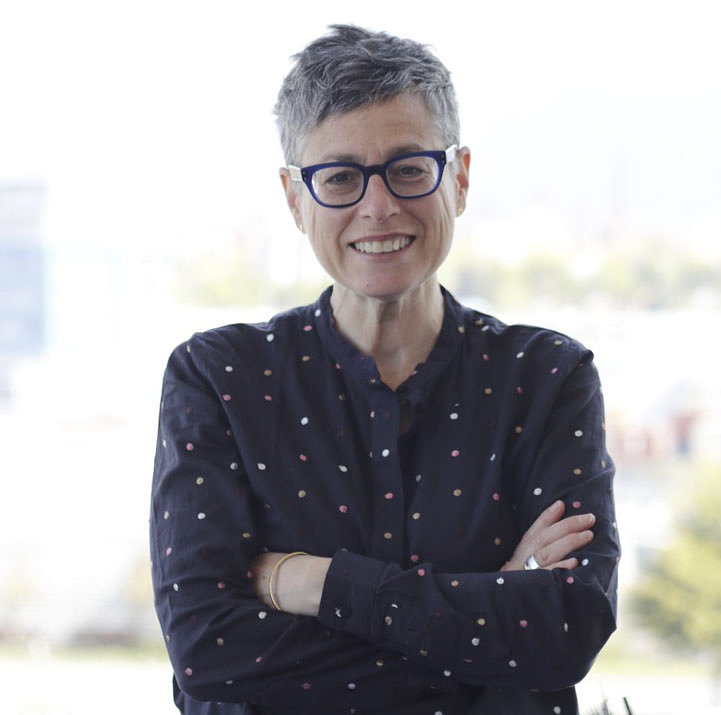 Randy Lee Cutler smiles with her arms crossed. She has short silver hair, and wears black glasses and a black button-up shirt with colourful polka dots.