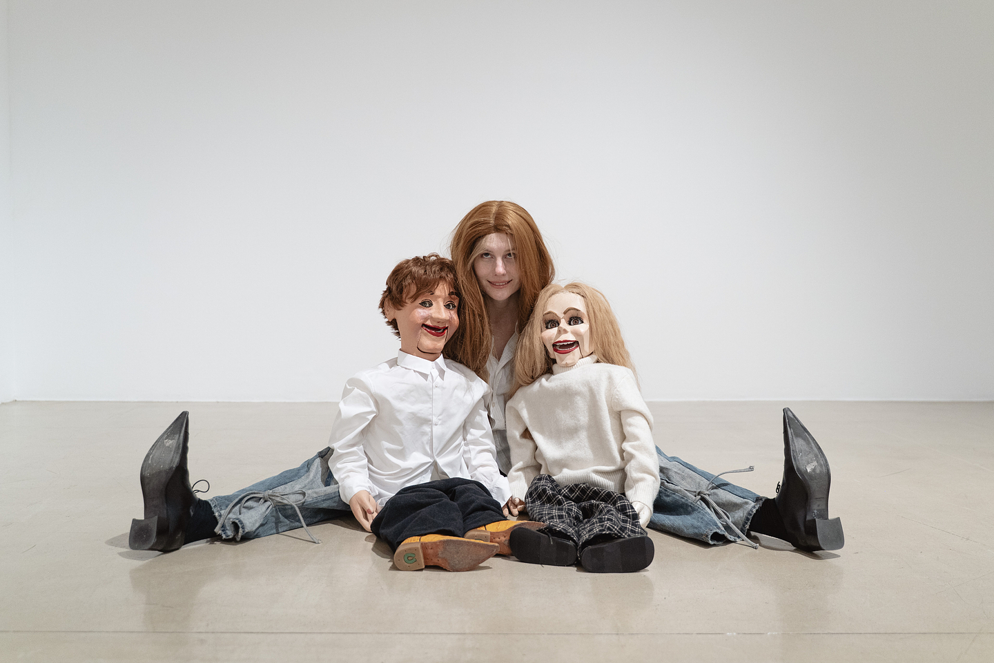A person with a long brown wig, white collared shirt, blue jeans, and black pointy toed shoes sits on a gallery floor with their legs spread wide, holding two ventriloquist puppets. One has short tousled brown hair and the other has long blonde hair. They are both dressed in white shirts. 
