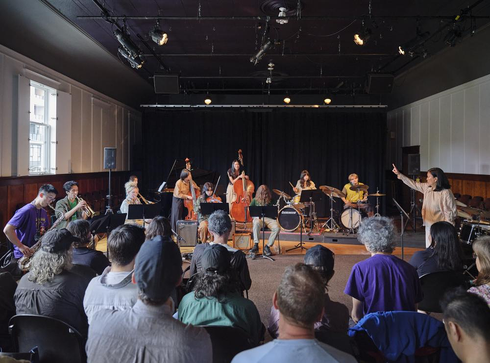 An audience, seen from behind, watches The New Improvisers play in the Grand Luxe Hall. On the right, Susie Ibarra conducts the performers. She has one arm raised just above her shoulder, with her index finger pointing towards the ceiling.