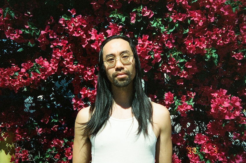 Patrick Shiroishi stands outdoors in front of a dense wall of deep red and magenta bougainvillea flowers. He has long dark hair, round wire-framed glasses, and wears a white ribbed tank top. The photo has a warm, film-like quality with dappled sunlight.