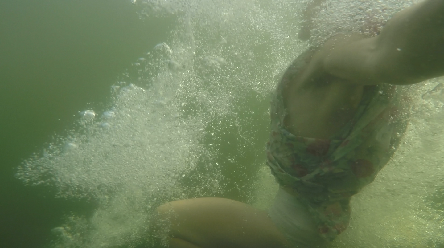 Bubbles dynamically swirl around a person submerged in a green-hued body of water. Their sleeveless floral sundress bunches up around their waist as they tread.