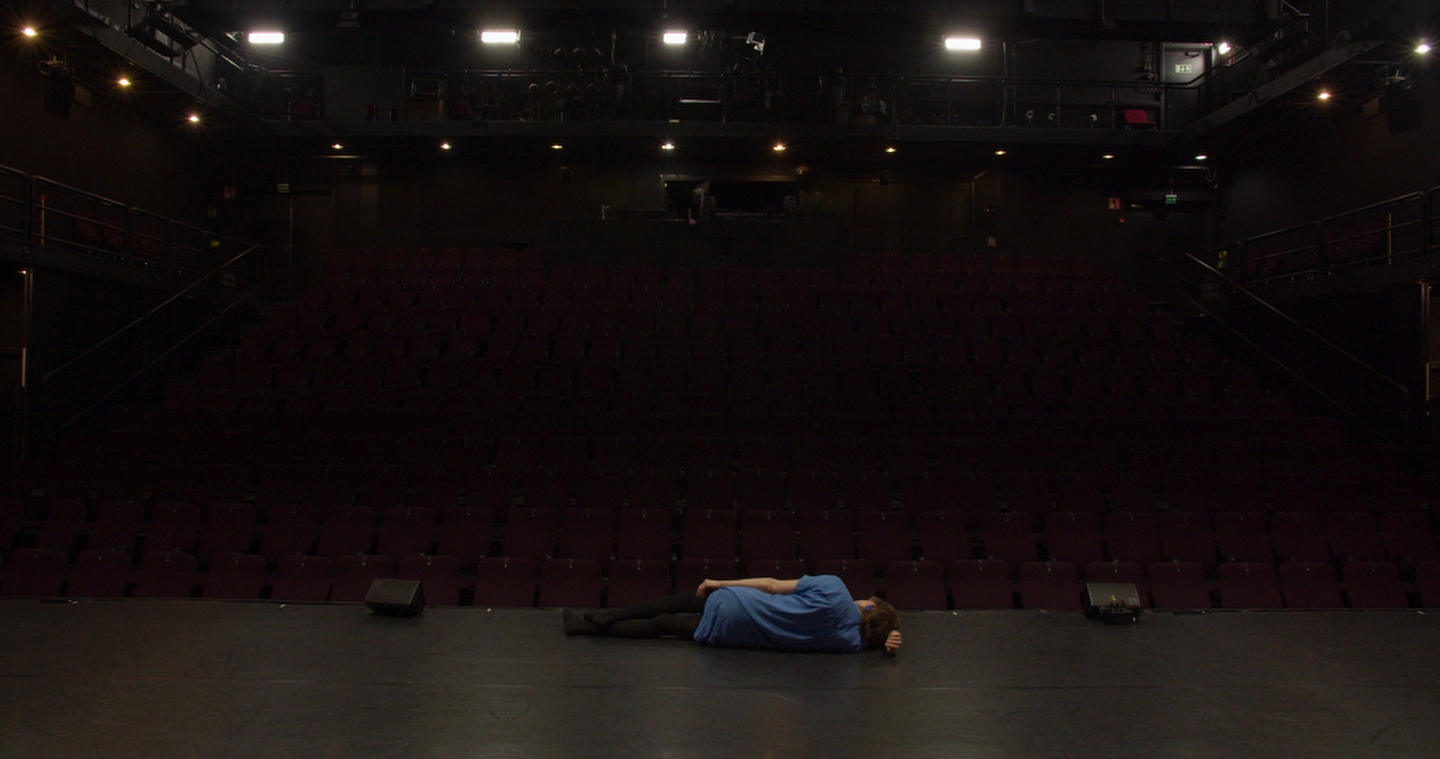 A person wearing a blue shirt lies curled up on an empty theater stage, facing away from the camera. The auditorium is dark and empty, with rows of unoccupied maroon seats and stage lights casting a dim glow.