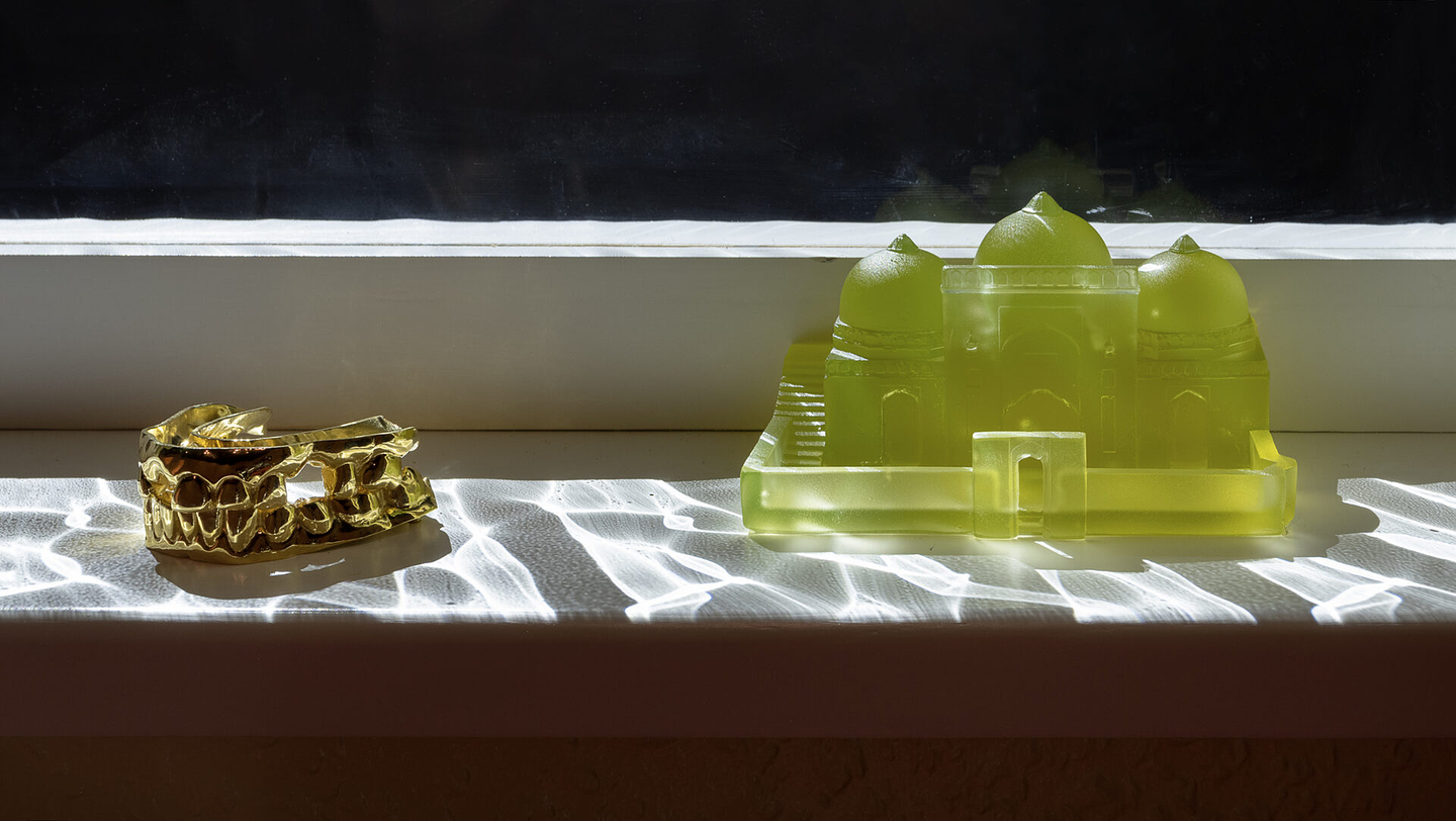 Two small sculptures sit side by side on a sunlit white windowsill. On the left is a shiny gold cast of a set of teeth. On the right is a translucent yellow-green model of a domed building with three domes and arched doorways inside a low walled enclosure. Rippling light patterns, like reflections off water, move across the sill.
