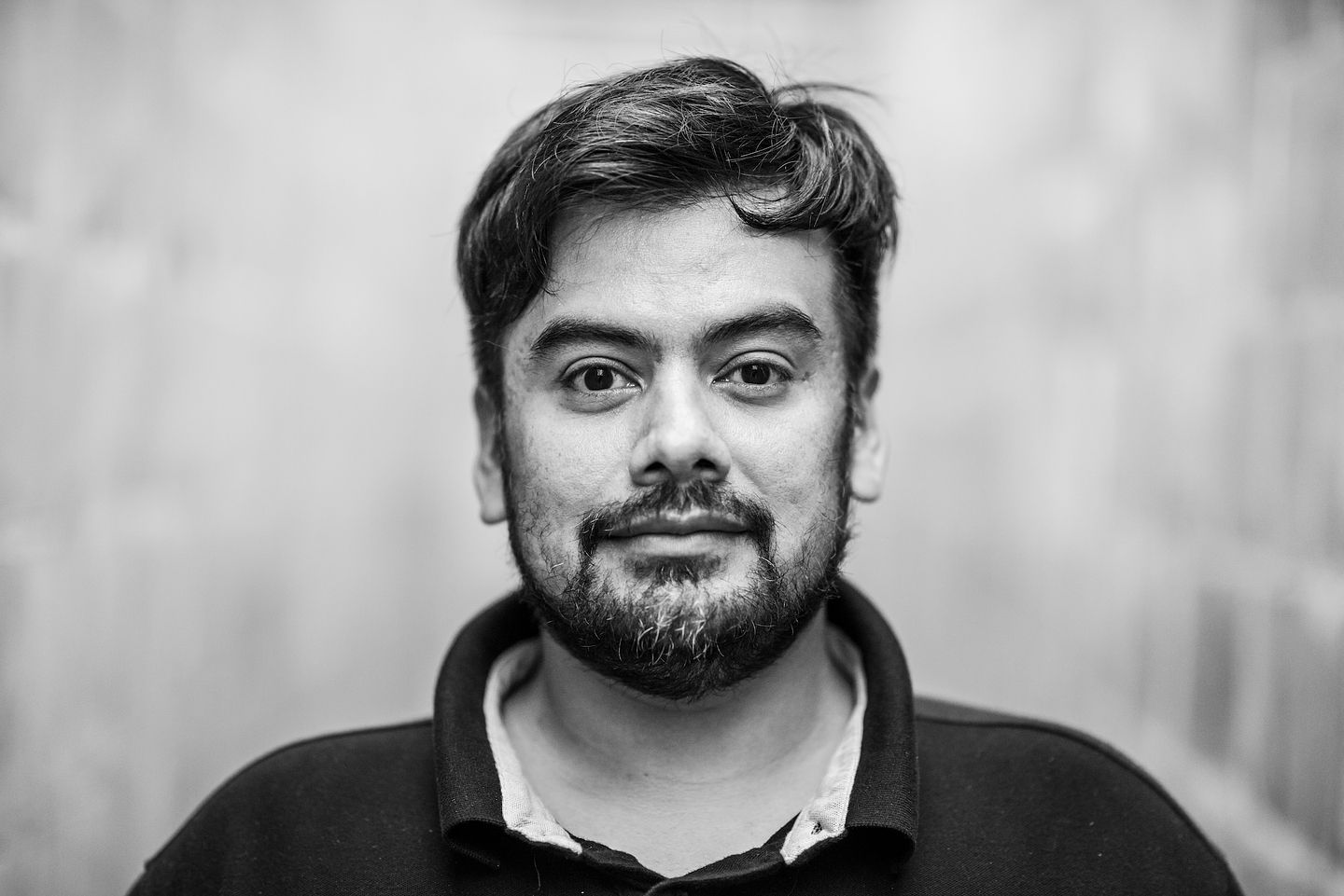 Black-and-white portrait of Naufus Ramírez-Figueroa with tousled dark hair and a beard, wearing a dark collared shirt over a light undershirt and looking directly at the camera with a slight smile. The background is a soft, out-of-focus pale wall.