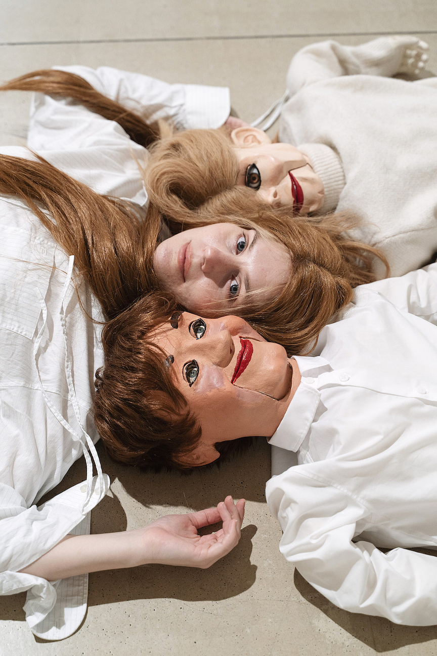 An overhead shot of a person with a long brown wig lying on the floor sandwiched between two ventriloquist puppets, one with brown hair and the other with light blonde hair. They are all wearing white shirts. 
