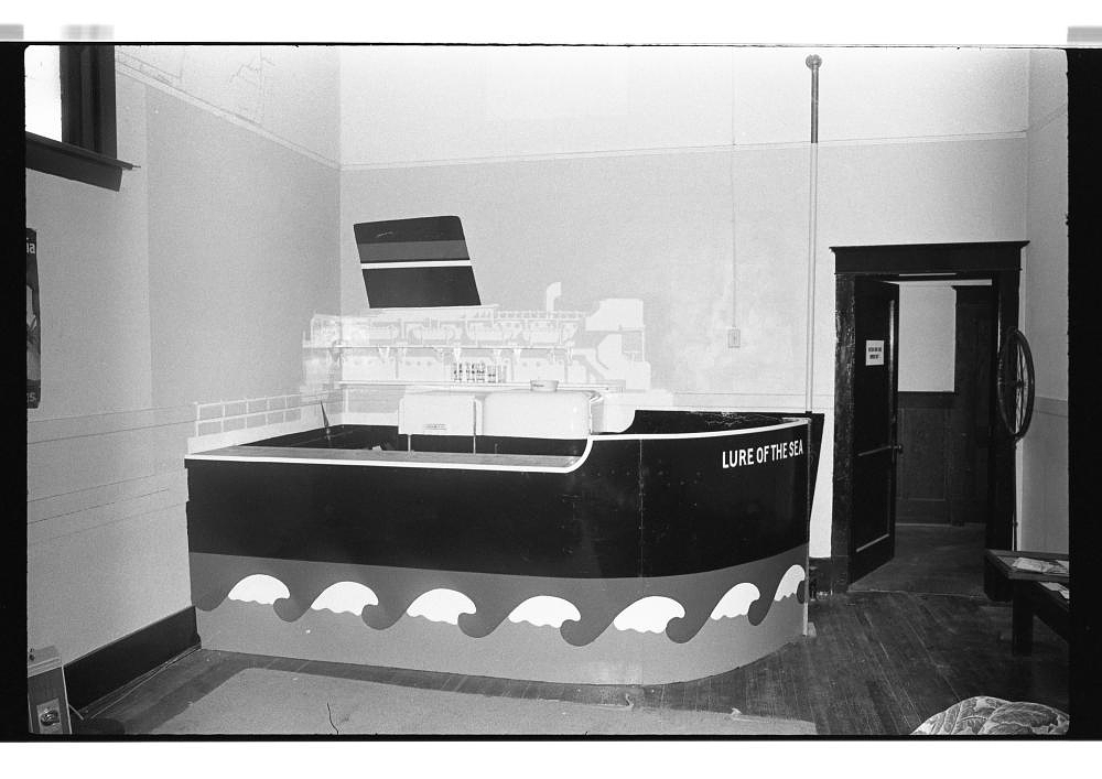Black-and-white archival photograph of an indoor gallery space featuring a bar modelled as a large cruise ship. The boat’s dark hull is decorated with stylized white waves and labelled “LURE OF THE SEA”; an open doorway, wooden floor, and a small bench appear in the surrounding room.