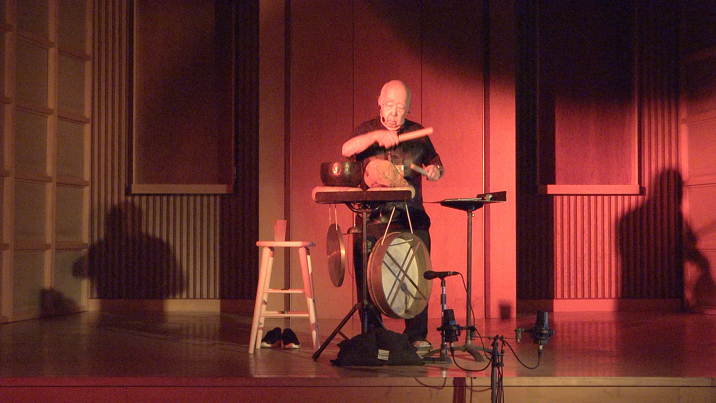 Nobuo Kubota stands on a stage in warm red lighting, playing percussion instruments arranged on stands, with a stool, microphone, and large drum nearby, and shadows cast on the wooden-paneled walls behind.