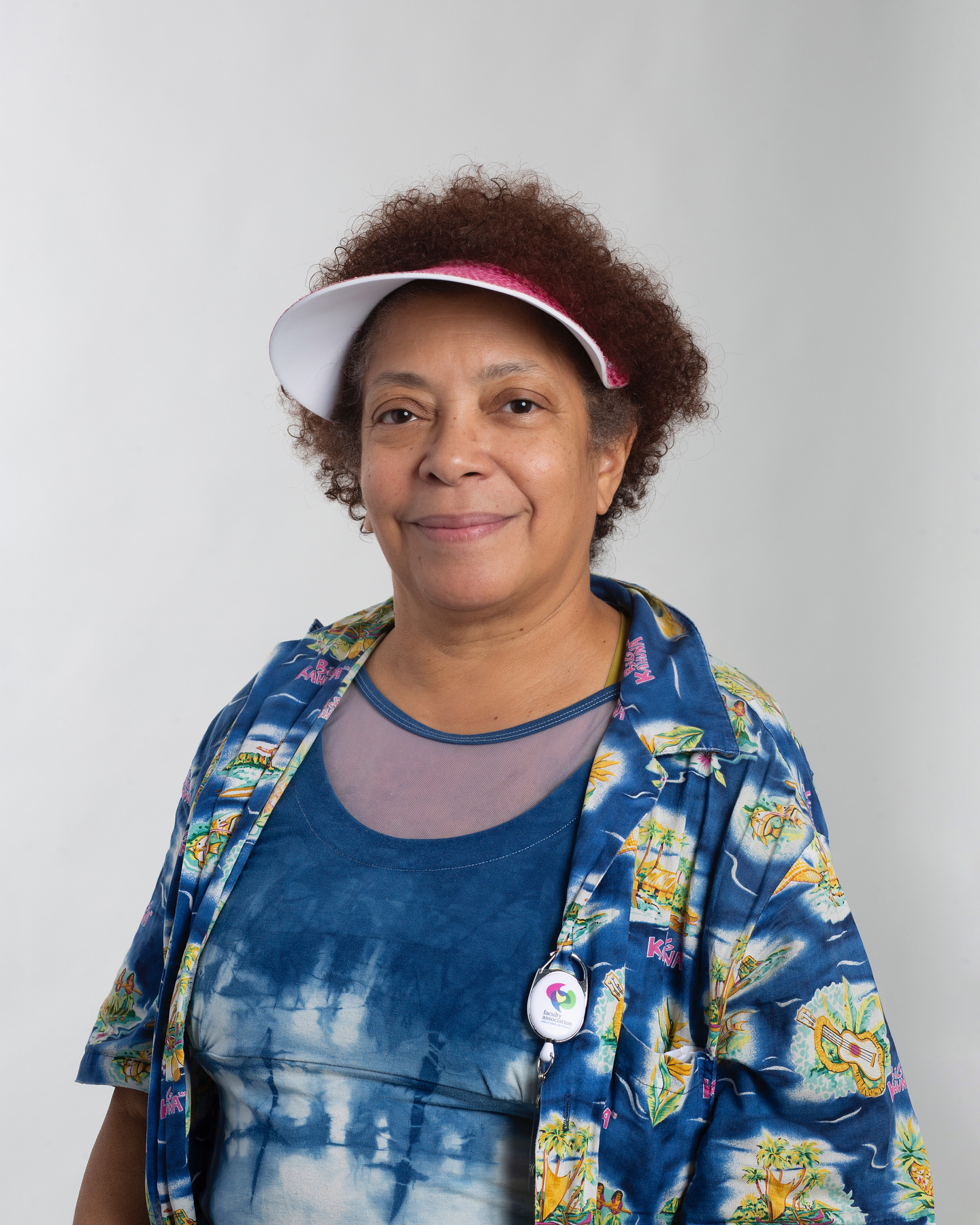 Smiling, Valerie Walker stands in front of a white background. She wears a short-sleeve shirt with Hawaiian iconography layered on top of an indigo dyed top. A hot pink visor peeks out from her short curly auburn hair. 