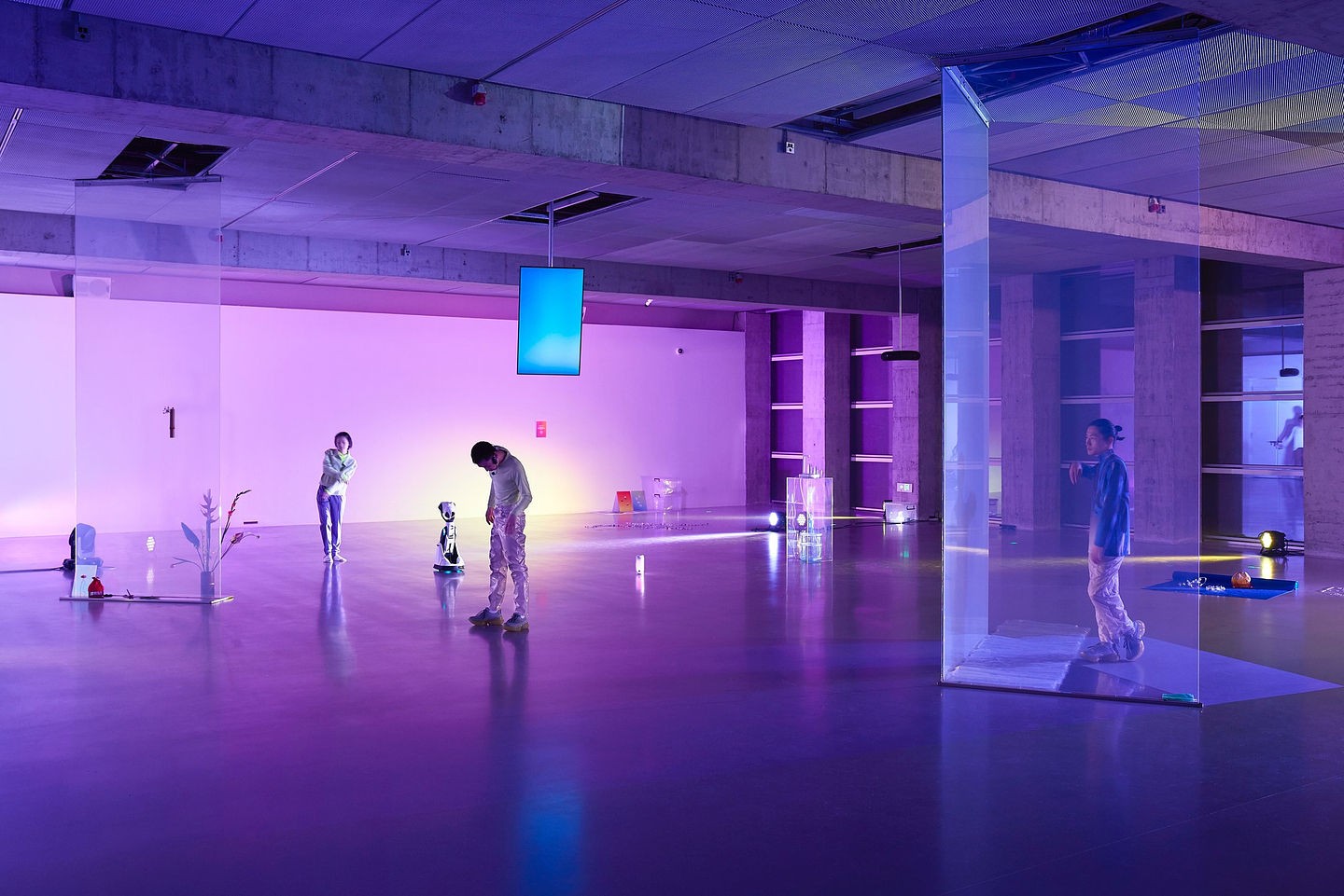 A photograph of an exhibition installed in a large open plan space. Two transparent screens are placed at either end of the space and reach from the floor to the ceiling, while a LED monitor suspended from the middle of the ceiling plays back a blue screen. A flower arrangement, small robot, clear perspex plinth, and other objects are placed intermittently at various points across the floor. In between the objects three performers can be seen enacting simple gestural movements. The space is lit by vibrant purple lights.