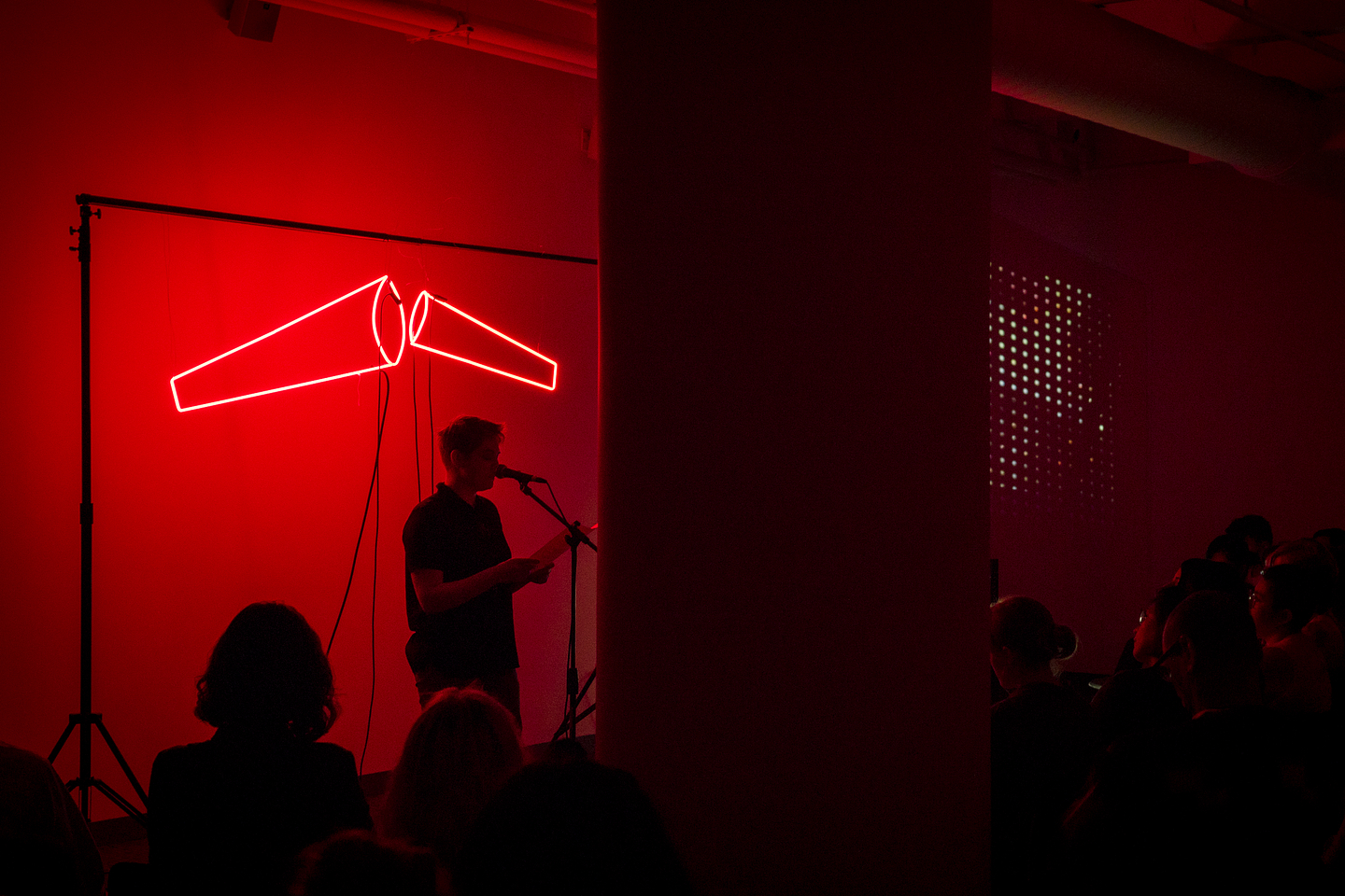 A person with short hair reads at a microphone for a seated audience in a gallery space. They are backlit by a red neon sign that resembles two megaphones.
