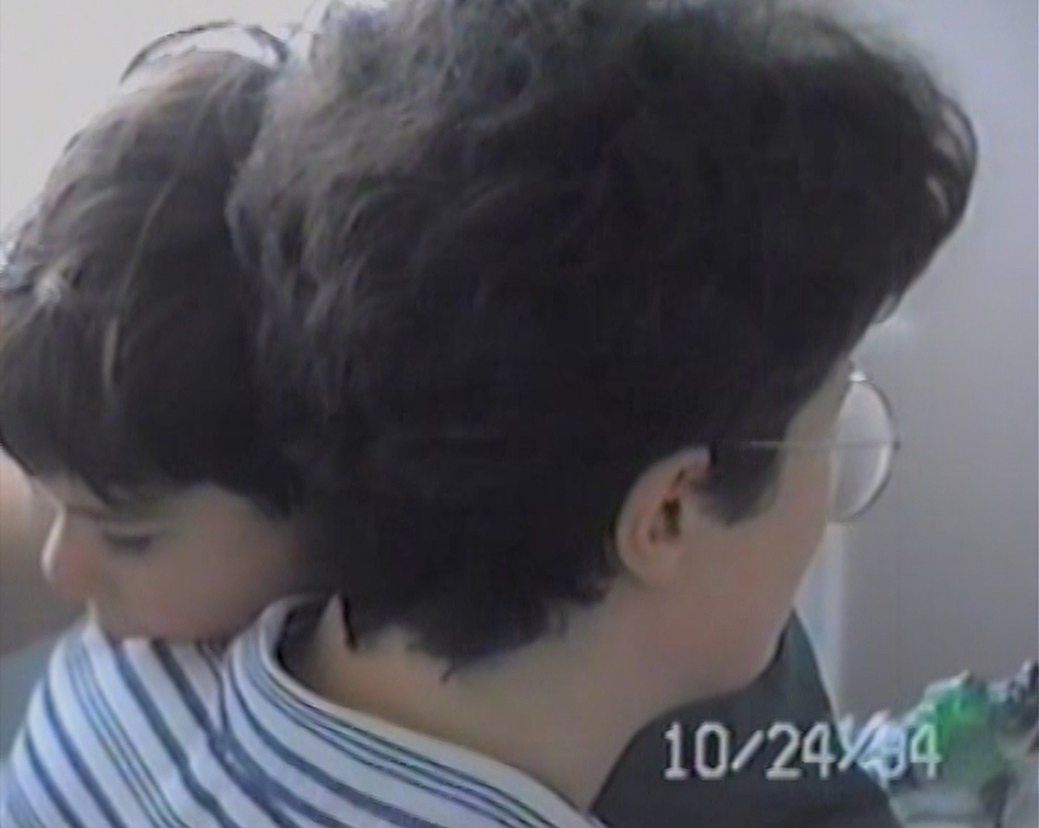 A camcorder still showing a close-up of brown-haired child draping their head over a parent's shoulder. The parent, who also has short brown hair, faces away from the camera and wears glasses and a striped collared shirt. The date October 24 1994 appears in the bottom right corner. 