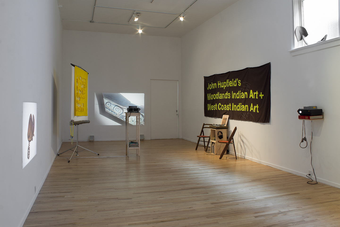 A colour photograph of the interior of Western Front’s gallery and Maria Hupfield’s installation. The exhibition consists of projected images on the north and west walls, a large black banner with yellow type that reads “John Hupfield’s Woodlands Indian Art + West Coast Indian Art” on the east wall, a small podium under the banner upon which sits a turnable, with a collection of records and two folding chairs next to it; and a sculpture that looks like a shovel with the handle bent at a right angle sitting on the windowsill high on the east wall.