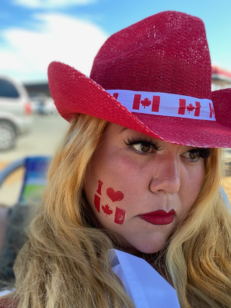 Close-up portrait of Skeena Reece, with long blonde hair, false lashes and red lipstick, gazing sideways toward the camera with a serious expression. She wears a red woven cowboy hat with a Canadian flag ribbon band, and her cheek has a temporary tattoo reading "I ♥" over a Canadian flag. Behind her, out of focus, are a blue sky and a parking lot.