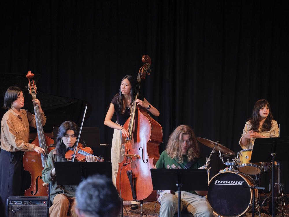 At the centre of the stage in the Grand Luxe Hall, five musicians play their instruments. Two stand, playing double basses. The others sit, one with a violin, another with a bass guitar, and the last in front of a drum set.