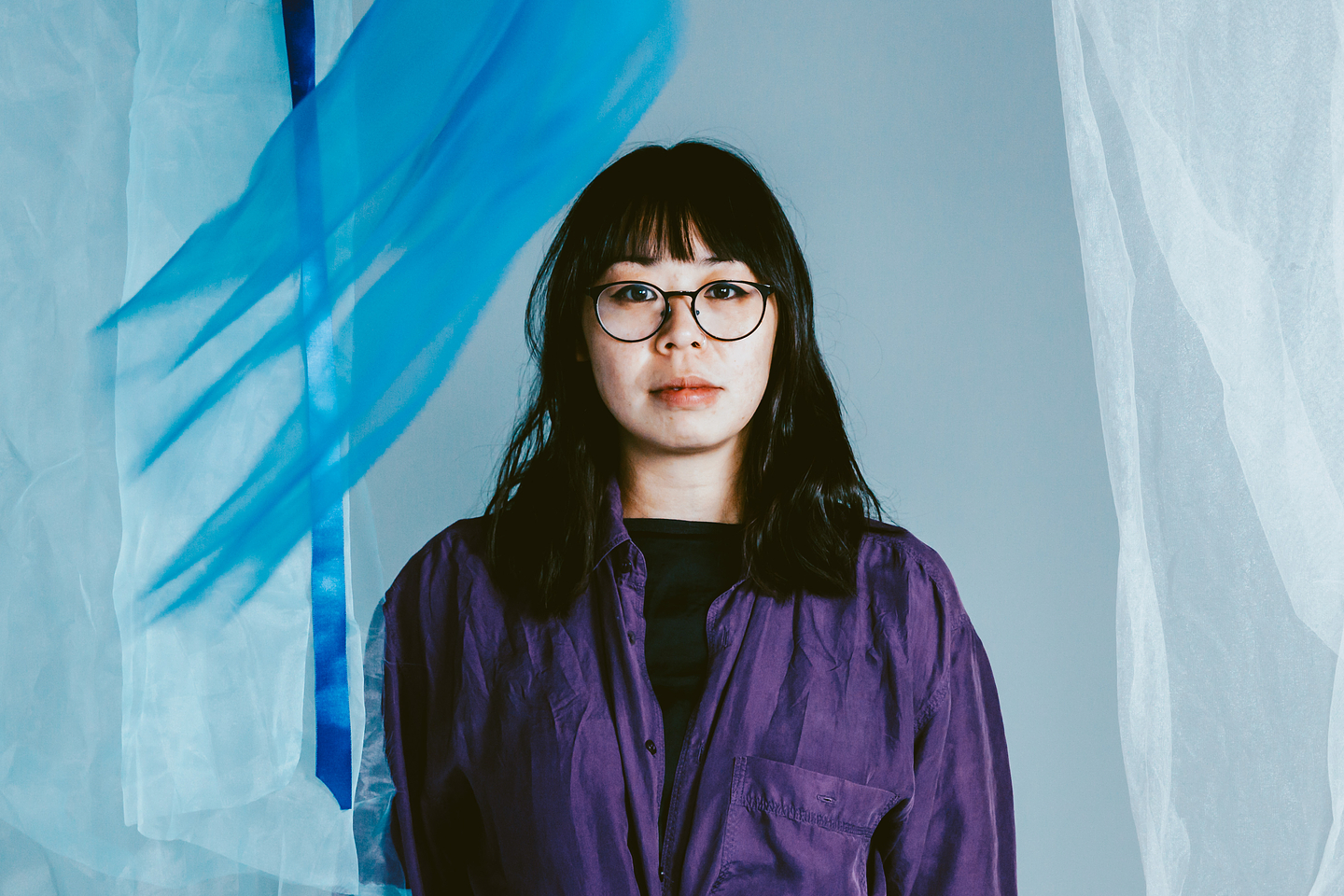 Karen Ng, with glasses and shoulder-length dark hair, faces the camera against a light blue background. She wears a loose purple button-up shirt over a black top and is partially framed by semi-transparent white and blue flowing fabric.