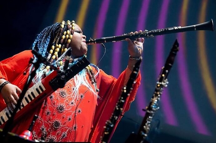 Angel Bat Dawid wears a bright red, patterned outfit and colourful braided hair while playing a clarinet on stage. She also holds a small keyboard instrument. Additional wind instruments are visible in the foreground against a dark background with curved, multicolored light patterns.