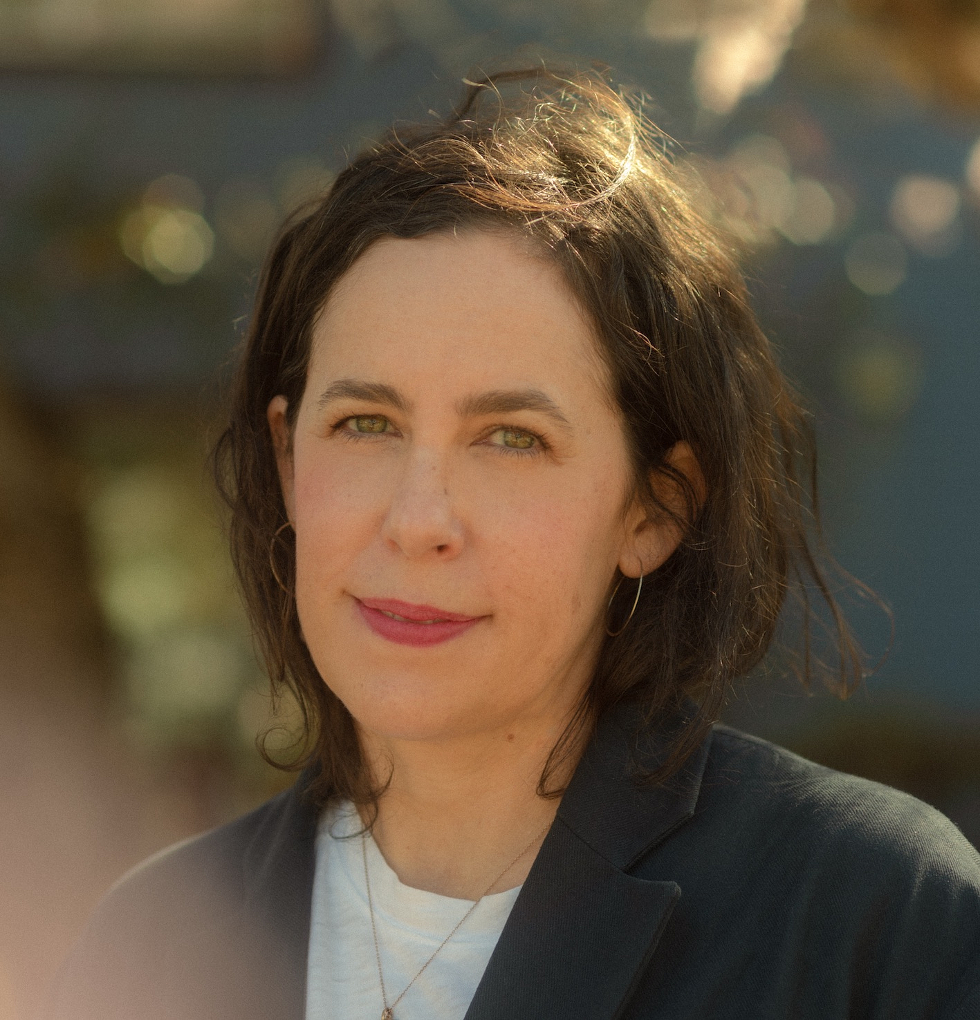A woman with dark hair and a gentle expression stands in soft sunlight, creating a serene, thoughtful mood.  She wears a black jacket and white top.