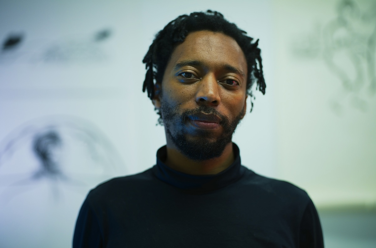 Portrait of Christian Nyampeta with short locs and a beard, wearing a black turtleneck and looking directly at the camera with a slight smile. Soft blue-green light falls across his face, and behind him, out of focus, are loose charcoal or ink drawings on a pale wall.