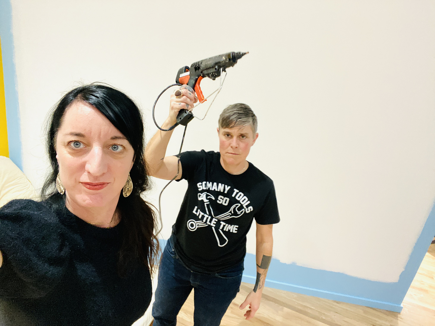 A selfie of Sheilah and Dani ReStack standing in front of a white wall that is in the process of being painted blue. Sheilah wears a black textured top and gold earrings. Dani, who is dressed in blue jeans and a black graphic t-shirt that reads "so many tools so little time," stands behind her while holding an industrial hot glue gun overhead. 