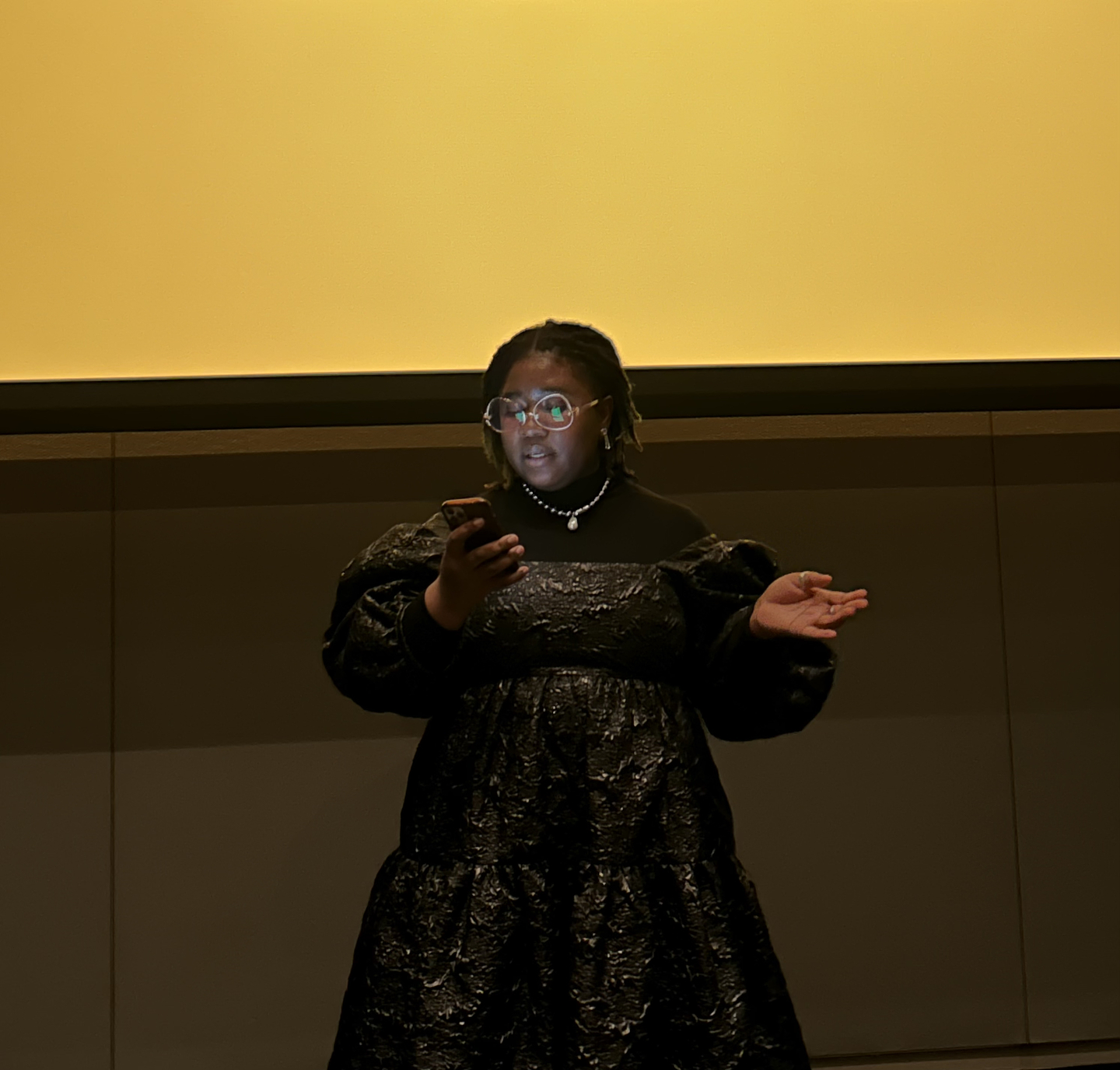 Wearing a black and silver puff-sleeved dress, Fegor Obuwoma stands in front of a yellow and grey wall while reading off their phone. The light from the screen is reflected in their round wire-framed glasses, casting their face in a soft glow. 