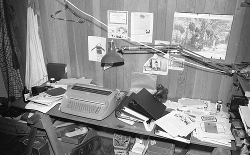 Black and white photo of a cluttered desk in a wood-paneled room, featuring a vintage typewriter, various papers, books, and folders scattered across the surface. A desk lamp extends over the workspace, and posters and drawings are pinned on the wooden wall behind the desk. A coat and hanging garment are visible to the left.