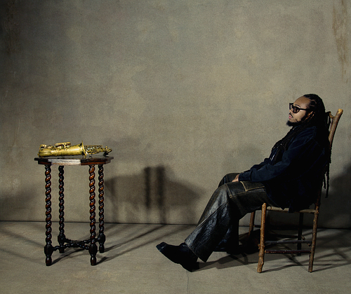Immanuel Wilkins wears sunglasses as he sits in a wooden chair facing left in a minimalistic, dimly lit room. He wears a dark jacket and leather pants. To his left is a small ornate wooden table with a brass saxophone resting on it. The background is a plain, textured wall, casting soft shadows.